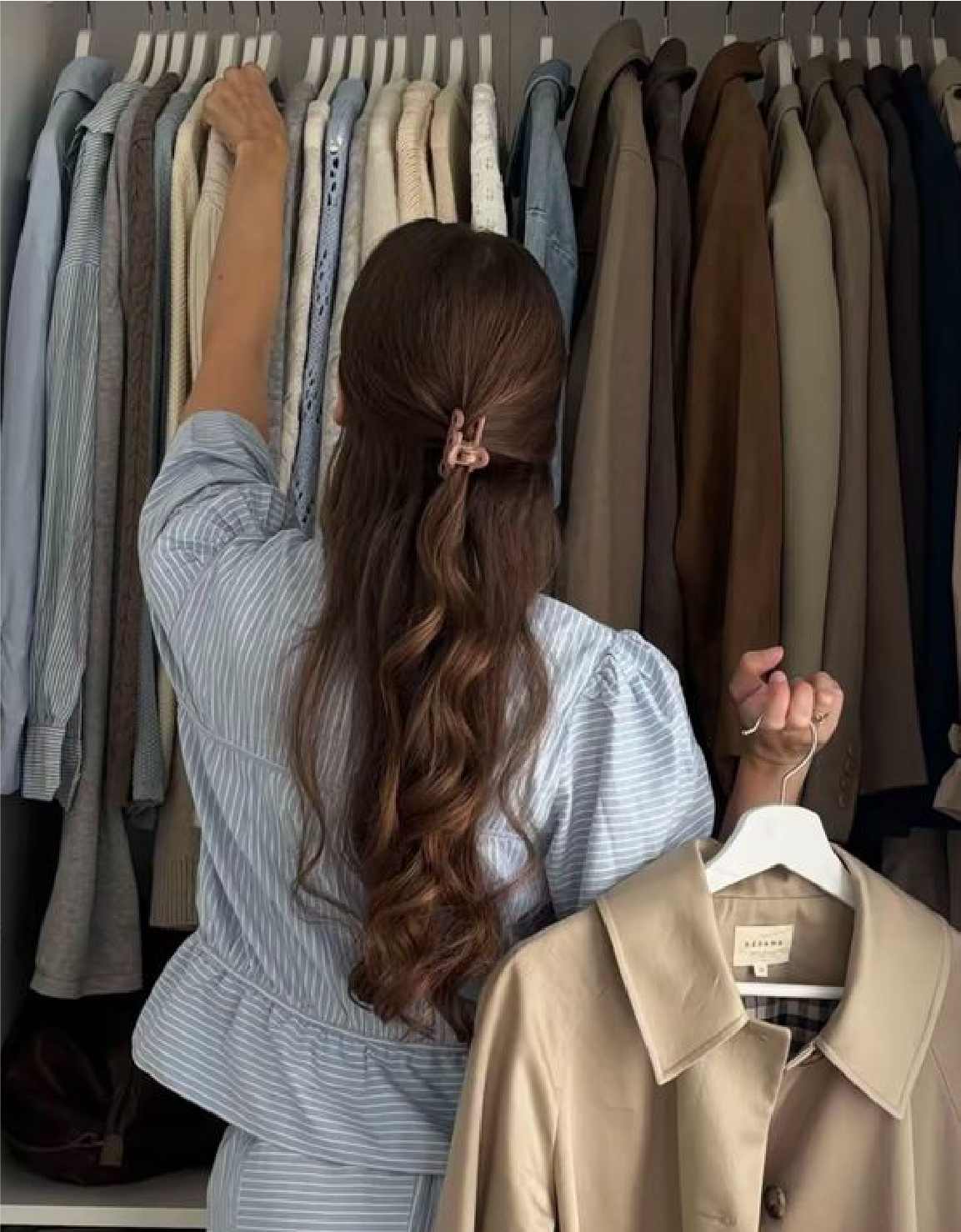 Mujer mirando ropa en un armario con prendas colgadas. Estilo relajado y moderno con chaqueta beige en la mano.