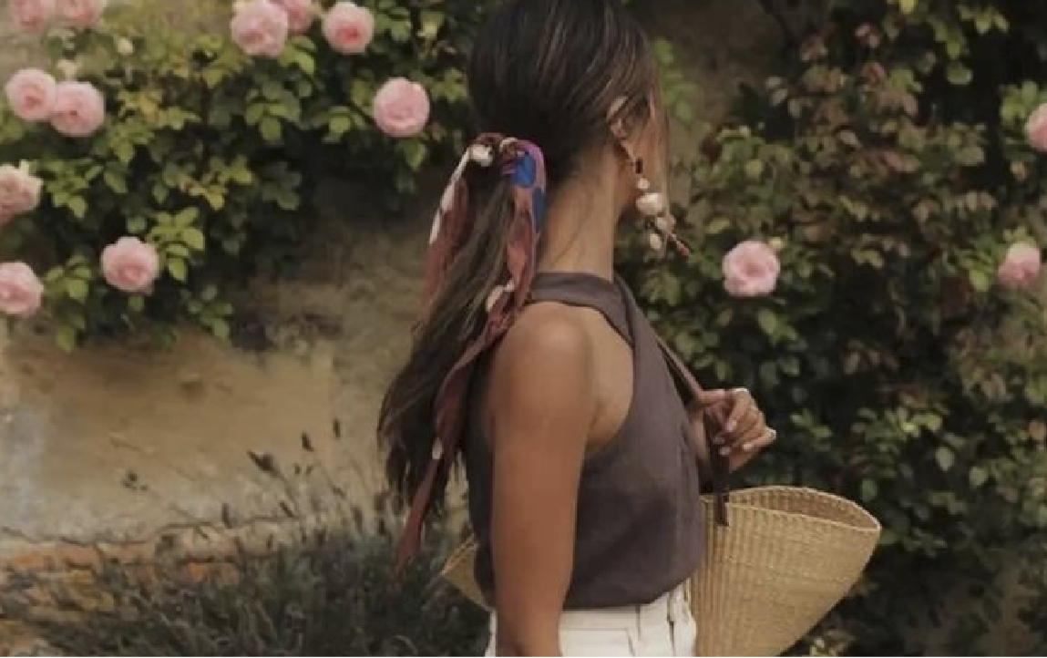 Mujer de espalda con top marrón, pañuelo en el pelo y bolso de mimbre junto a rosales de Marfranc